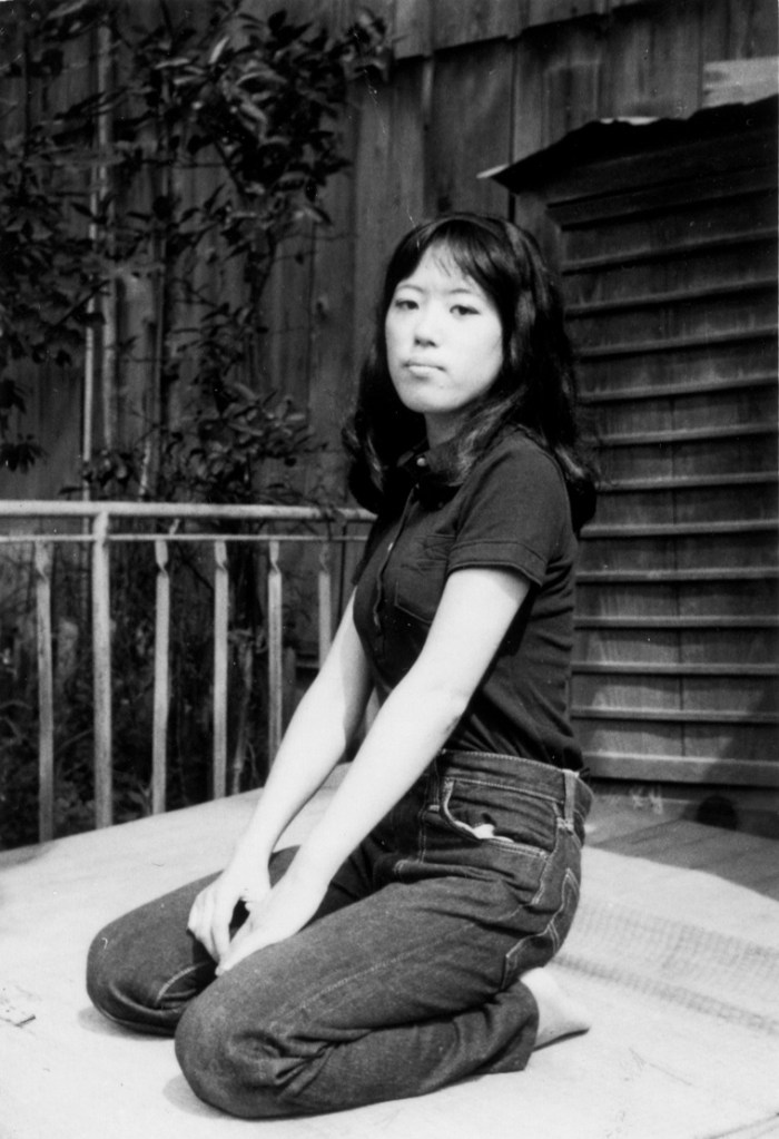 Black and white photograph of a young Asian woman kneeling on the ground, with her legs tucked fully underneath her. Her left side is angled towards the camera. She has shoulder length dark hair, and is wearing a dark t-shirt and jeans. Her hands are resting on her lap. She appears to be outside. There is a light fence to her right, and shuttered metal door behind her. /end