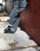 Photograph of the lower half of a person outside, leaning against a red brick wall. Only their legs and the lower half of their stomach are visible. They are stood with their left side facing the camera. They are wearing blue jeans with a long metal chain next to their left hip, and black boots. Their feet are crossed at the ankle. /end