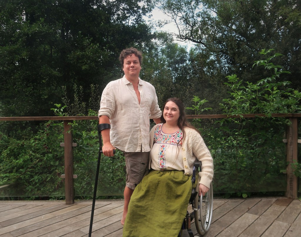 Photograph of two people on a wooden deck outside, in front of a lot of green trees. The man on the left is standing. He is white, has curly brown hair and is wearing a loose white shirt and brown shorts. His right leg is amputated below the hip. He has a black forearm crutch on his right arm. His left arm is partially obscured by the woman next to him. She is sitting in a manual wheelchair. She is white, has long brown hair and is wearing a long-sleeved white cardigan over a floral blouse, and a long green skirt. They are both smiling. /end