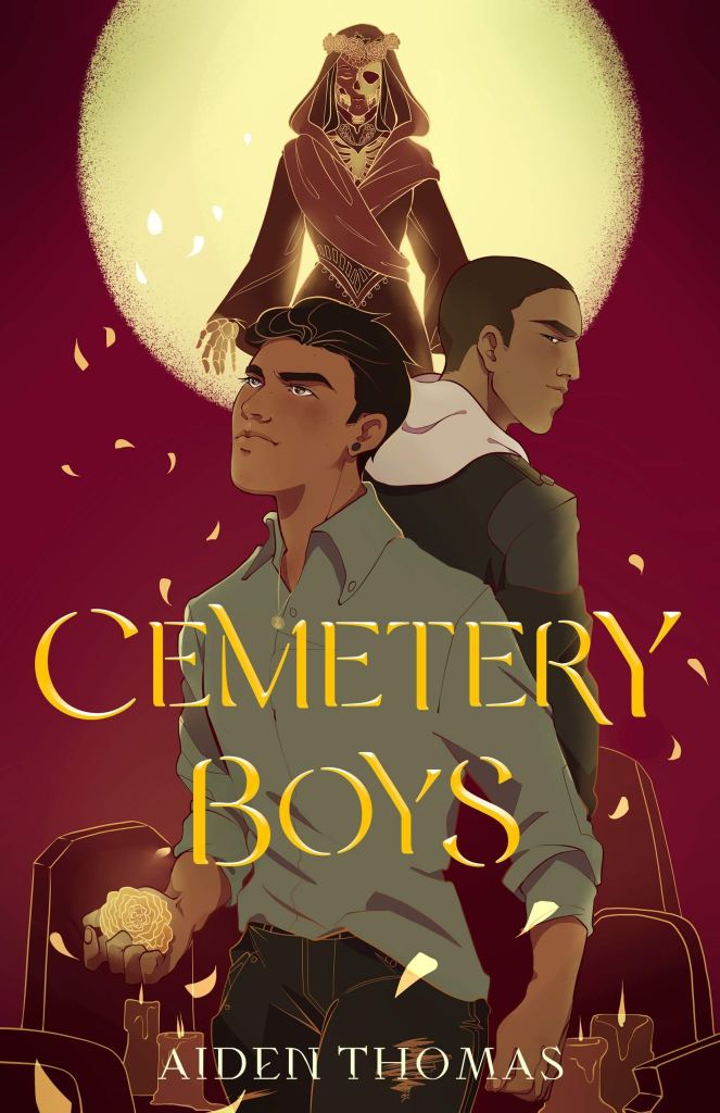 A book cover. The title "Cemetery Boys" is written in the lower centre of the cover, in large gold capitals. The name of the author "Aiden Thomas" is written at the bottom in small white capitals. The background is red and shows two men in front of gravestones, under a large full moon. The man in the foreground is wearing a grey long-sleeved shirt, unbuttoned at the collar, and black pants. He has a yellow marigold in his right hand. The man behind him has short dark hair and is wearing a dark jacket with a white hood. Behind them both, in the centre of the cover, at the top, a skeletal figure in a long red robe with a red hood and yellow marigold crown. /end
