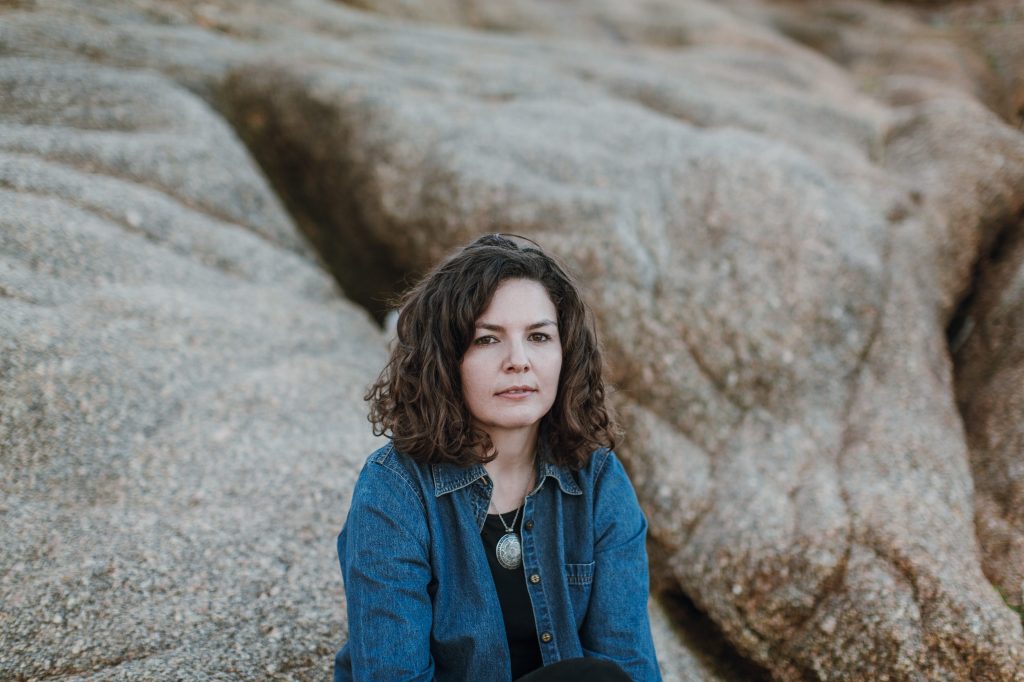 Photograph of a white woman sitting on large rocks outside. She has short bobbed, wavy brown hair. She is wearing an open denim shirt over a black t-shirt and has a necklace with a large circular pendant around her neck. /end
