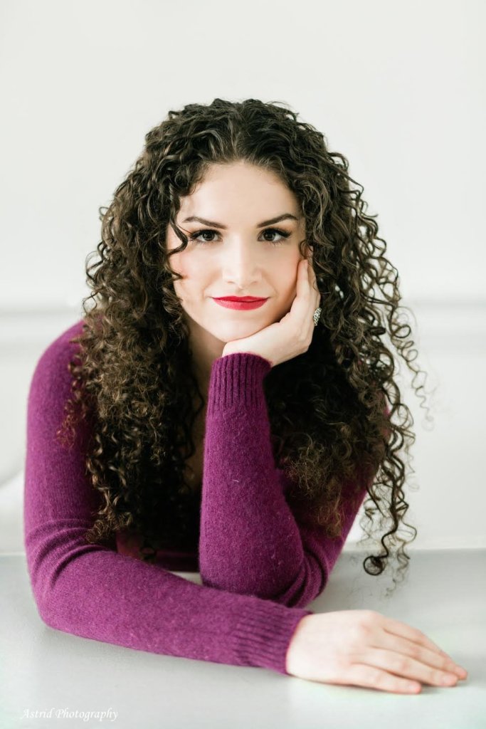 A white woman with brown eyes, wearing a purple sweater, facing the camera. She has long curly brown hair and her cheek rests on her left hand. /end