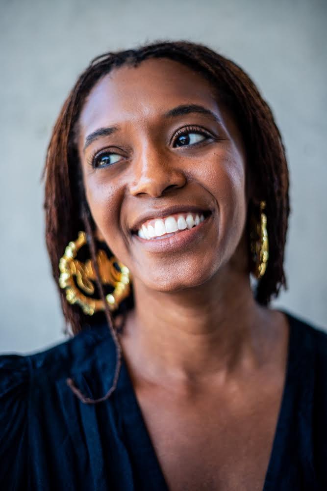 Photograph of a black person against a light grey background. They are smiling widely and looking off to the left side of the image. They have long brown hair, with a small thick strand resting on their right shoulder. They are wearing a deep v-neck, navy blue blouse and large circular golden earrings with an unidentifiable shape in the centre. /end