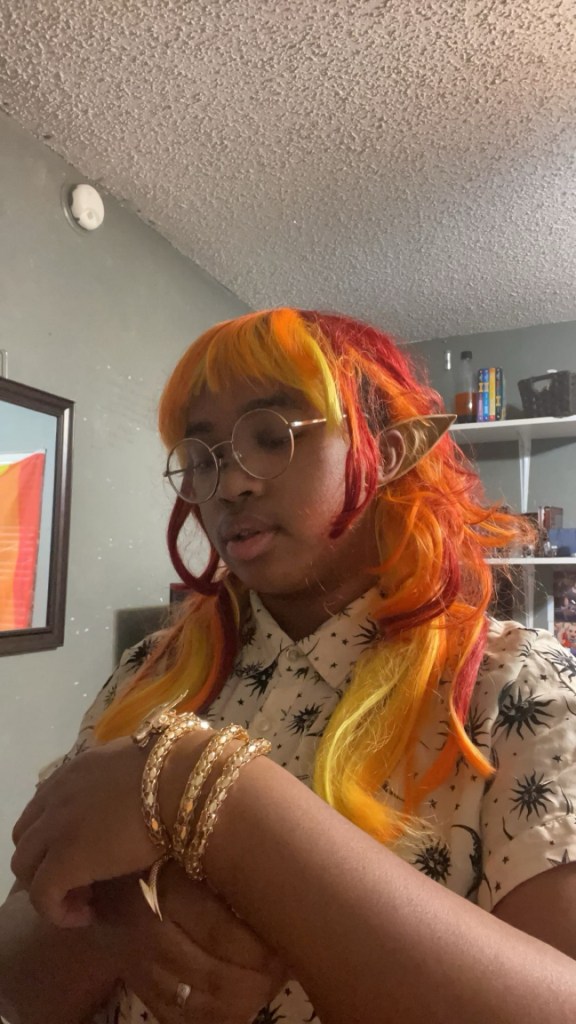 Photograph of a black person wearing a long orange, red, and yellow wig, and pointed ears. They are wearing round gold glasses and a white shirt with black suns, moons and stars across it. On their left wrist there is a golden bracelet made to look like a dragon wrapping around the arm. There is a white shelving unit behind them, and a mirror to their right with a partial reflection of the progress pride flag. /end