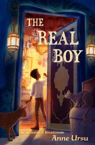 Cover for the book 'The Real Boy' by Anne Ursu. The title is written at the top in large white capitals. The author's name is written in the lower right corner in smaller yellow capitals. Cover art shows a young boy standing in the entry way to a bright, cluttered room. Behind him, a darker, blue room, with two cats. There is a large ornate door to his right. /end