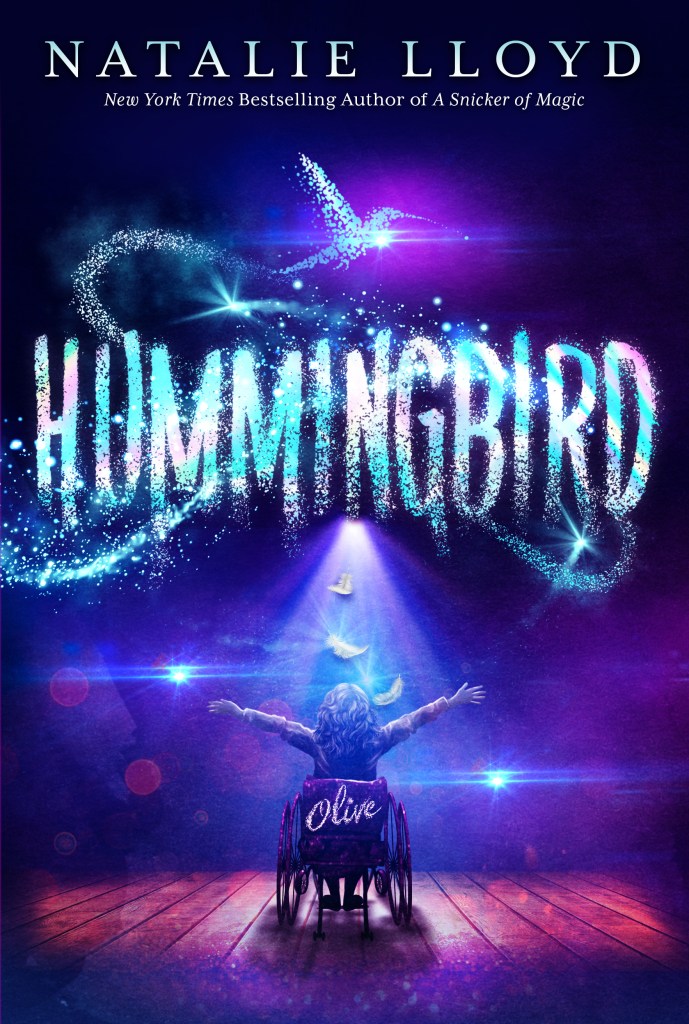 Cover for the book "Hummingbird" by Natalie Lloyd. The authors name is at the top of the cove in white capitals. The title is in the centre of the cover, in large blue, pink and white glittering capitals. Cover art shows a person in a wheelchair with long hair. The wheelchair says "Olive" on the back. The person appears to be on a wooden floor or stage, with lights or an audience in front of them. There is a glittering blue hummingbird above the title. /end